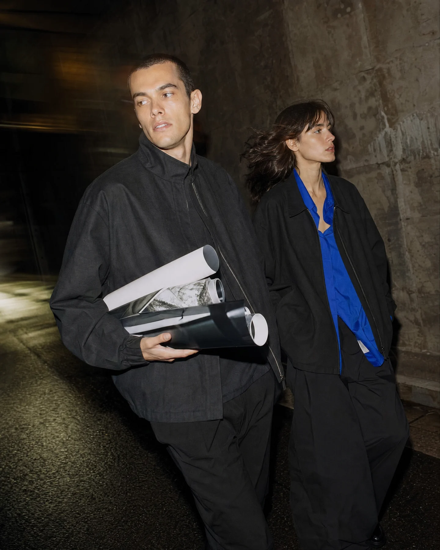Two adult creatives in unbranded streetwear walking through a nighttime city alley under editorial flash.